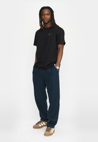 Zwart T-shirt met korte mouwen en een klein embleem, gecombineerd met marineblauwe broeken. Schoenen zijn tan kleurig met witte strepen. Het model staat met de handen in de zakken.