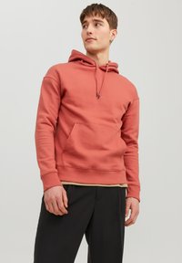 Roestkleurige hoodie van zacht materiaal, met een kangoeroezak, trekkoorden voor de capuchon en ribgebreide manchetten en zoom. Gedragen over een beige shirt.