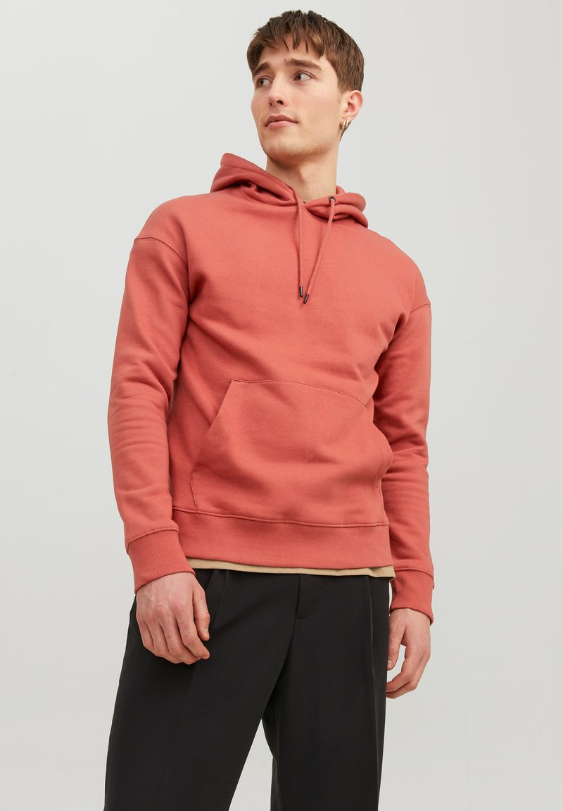 Roestkleurige hoodie van zacht materiaal, met een kangoeroezak, trekkoorden voor de capuchon en ribgebreide manchetten en zoom. Gedragen over een beige shirt.