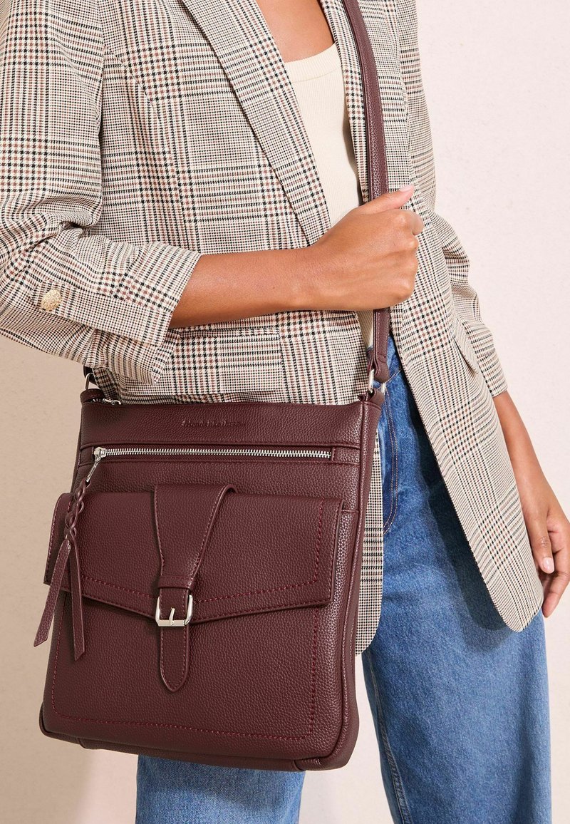 Žena drží tmavohnedú koženú crossbody tašku s predným vreckom na pracku, má na sebe károvaný blejzer a modré džínsy.