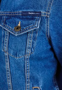 Kék farmerdzseki mellzsebbel, arany varrással és fekete gombbal. A mellzseb felett a "Pepe Jeans London" címke található. Sima textúrájú.