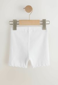 Short en tissu blanc côtelé avec ourlet festonné suspendu sur un cintre en bois avec pinces métalliques sur fond uni.