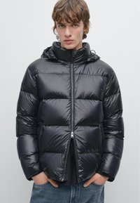 Zwarte pufferjack met een glanzende textuur, ritssluiting aan de voorkant en een capuchon. Bevat gevoerde secties en zijzakken. Gekozen over een donkere top, casual gestyled.