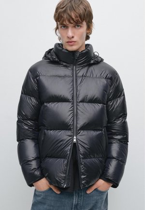 Schwarze Daunenjacke mit glänzender Oberfläche, Reißverschluss vorne und Kapuze. Verfügt über gepolsterte Einsätze und seitliche Taschen. Wird über einem dunklen Oberteil getragen, lässig gestylt.