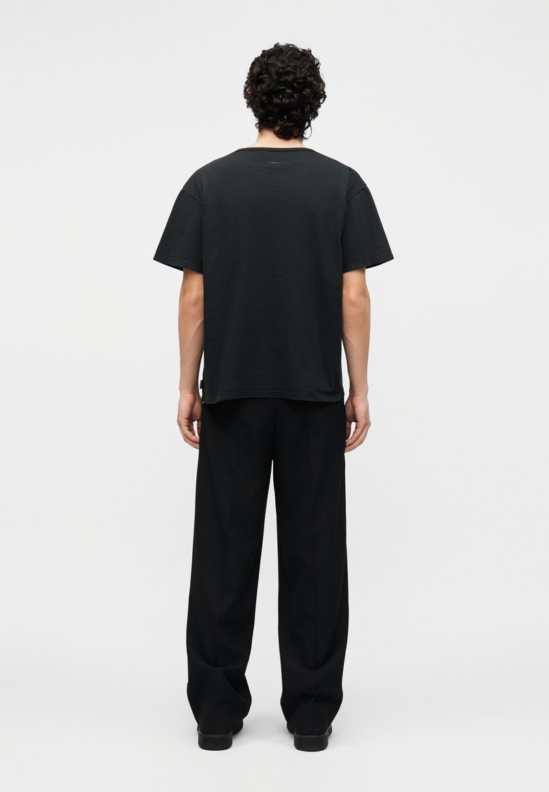 Homme aux cheveux bouclés, debout de dos, portant un t-shirt noir ample et un pantalon large noir sur un fond uni clair.