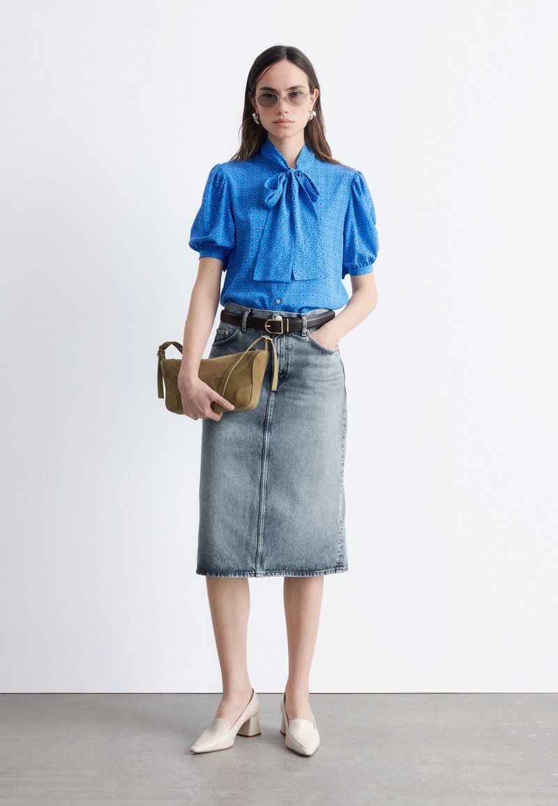 Femme portant un chemisier bleu à manches bouffantes avec un nœud, une jupe midi en denim délavé, des talons blocs beiges, des lunettes de soleil, et tenant une pochette beige.
