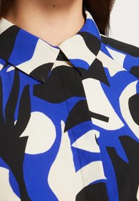 Košilová blůza s límcem, zdobená výrazným abstraktním vzorem v modré, černé a krémové barvě. Vyrobena z hladkého materiálu se strukturovaným designem.