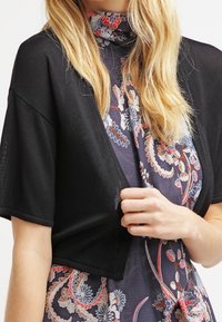 Svart kort cardigan med korta ärmar, tillverkad av lättstickat material, lagd över en blommönstrad klänning med blåa och röda mönster.