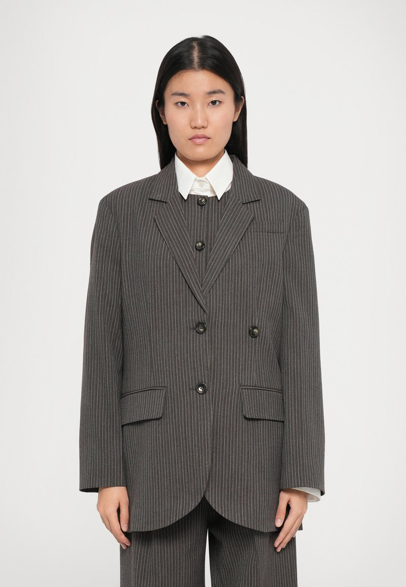 Siv pinstripiran blazer s prilagojenim krojem, dvorednim zapenjanjem, črnimi gumbi in zareznimi ovratniki. V kombinaciji z belo srajco s ovratnikom.