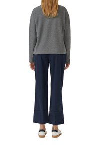 Personne avec de longs cheveux châtain clair, portant un haut à manches longues gris, un pantalon court large bleu marine et des baskets blanches, se tenant de dos.