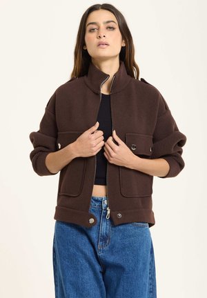 Brown fleece jacket with a stand collar, front zip, and large flap pockets. Features ribbed cuffs and open hem. Paired with blue jeans.