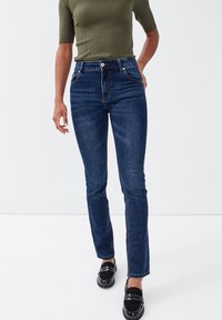 Hochgeschnittene, dunkelblaue Jeans mit Slim-Fit, vorderen Taschen und minimalen Steppdetails. Getragener strukturierter grüner Oberteil.