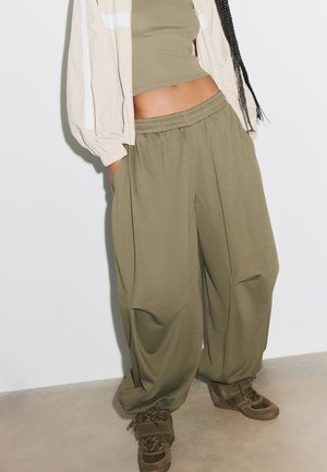 Personne portant un pantalon ample vert olive, un crop top assorti, une veste beige et des chaussures marron, debout contre un mur blanc.