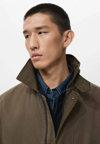 Chaqueta marrón con cuello alto, combinada con una camisa de mezclilla azul. Tejido suave y detalles de cremallera visibles. Presenta elementos de diseño moderno.