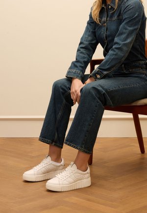 Witte sneakers met textuuraccenten, met een dikke zool en veters. Draagt ze met donkere denim broeken, zittend op een stoel met houten poten.