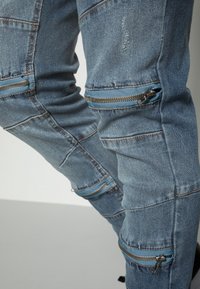 Ljusblå denimjeans med två fickor med dragkedja vid knäna och blå detaljer, samt ett sömnads mönster över tyget.