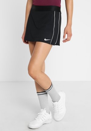 Person trägt schwarzen Nike-Sportrock mit weißen Streifen, graue Socken mit schwarzen Streifen und weiße Sportschuhe, steht mit überkreuzten Beinen.