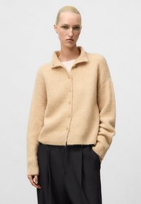 Cardigan en laine beige avec une texture douce, doté d'un col, d'une fermeture à bouton et d'un ourlet côtelé. Porté sur un pantalon large noir.