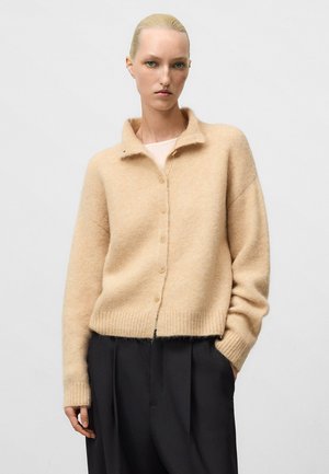 Beige wool cardigan with a soft texture, featuring a collar, button closure, and ribbed hem. Worn over black wide-leg trousers.