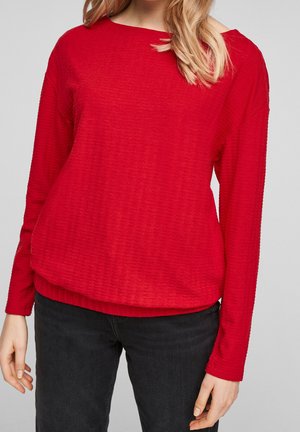 Kvinde iført en rød, tekstureret langærmet bluse og sorte bukser, stående mod en ensartet lys baggrund.