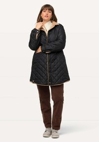 Quiltad svart kappa med tanfärgad krage, med tryckknappar, strukturärmar och en midjelång design. Bruna kordfläskbyxor och ljusa skor.