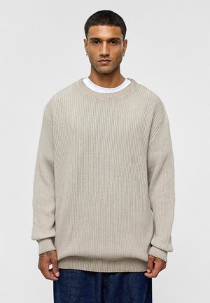 Beige gebreide trui met een ontspannen pasvorm, geribbelde textuur, ronde hals en lange mouwen. Draagt over een wit T-shirt en gecombineerd met donkere spijkerbroeken.