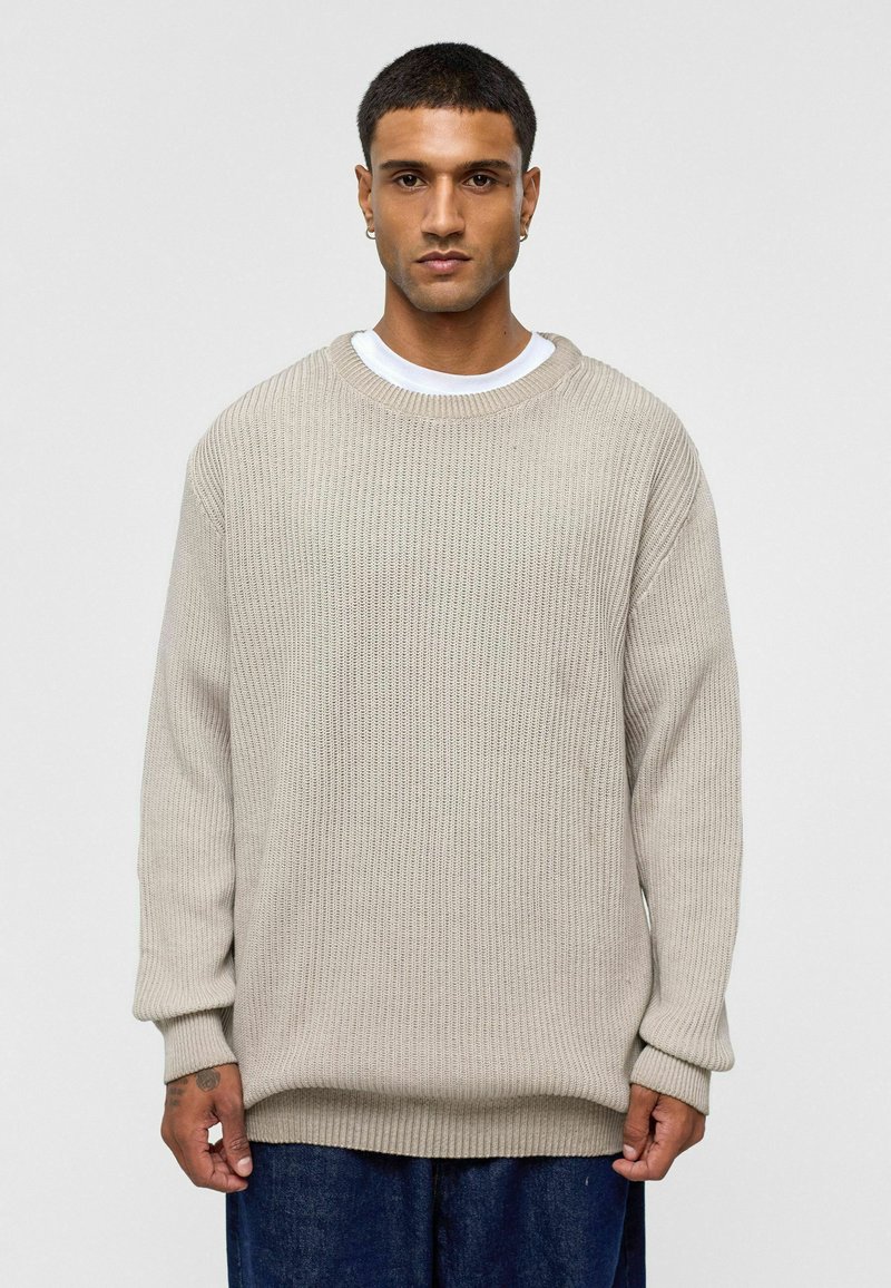 Beige gebreide trui met een ontspannen pasvorm, geribbelde textuur, ronde hals en lange mouwen. Draagt over een wit T-shirt en gecombineerd met donkere spijkerbroeken.