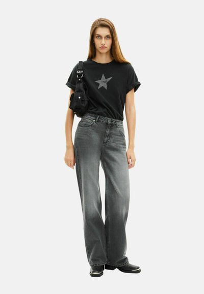 Camiseta negra con un gráfico de estrella gris, combinada con unos jeans grises de pierna ancha y alta cintura. Una pequeña bolsa negra está colgada de un hombro.