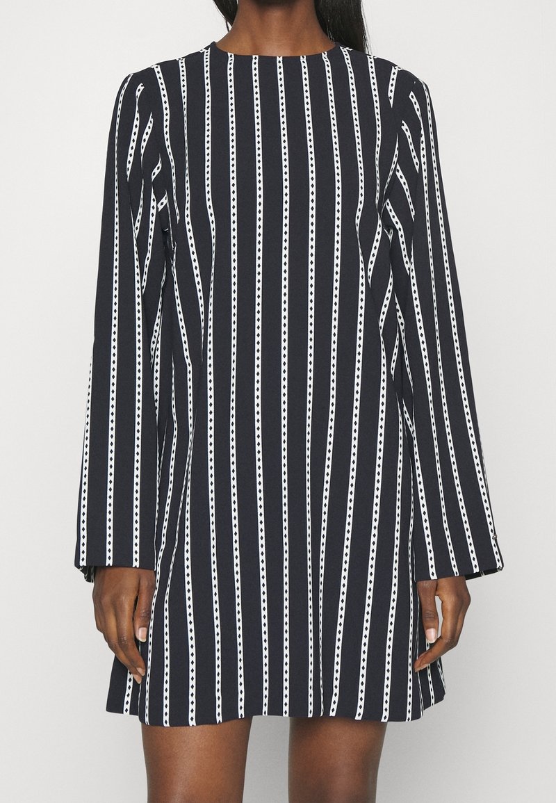 Femme portant une robe noire à rayures verticales blanches et manches longues, se tenant devant un fond blanc uni.