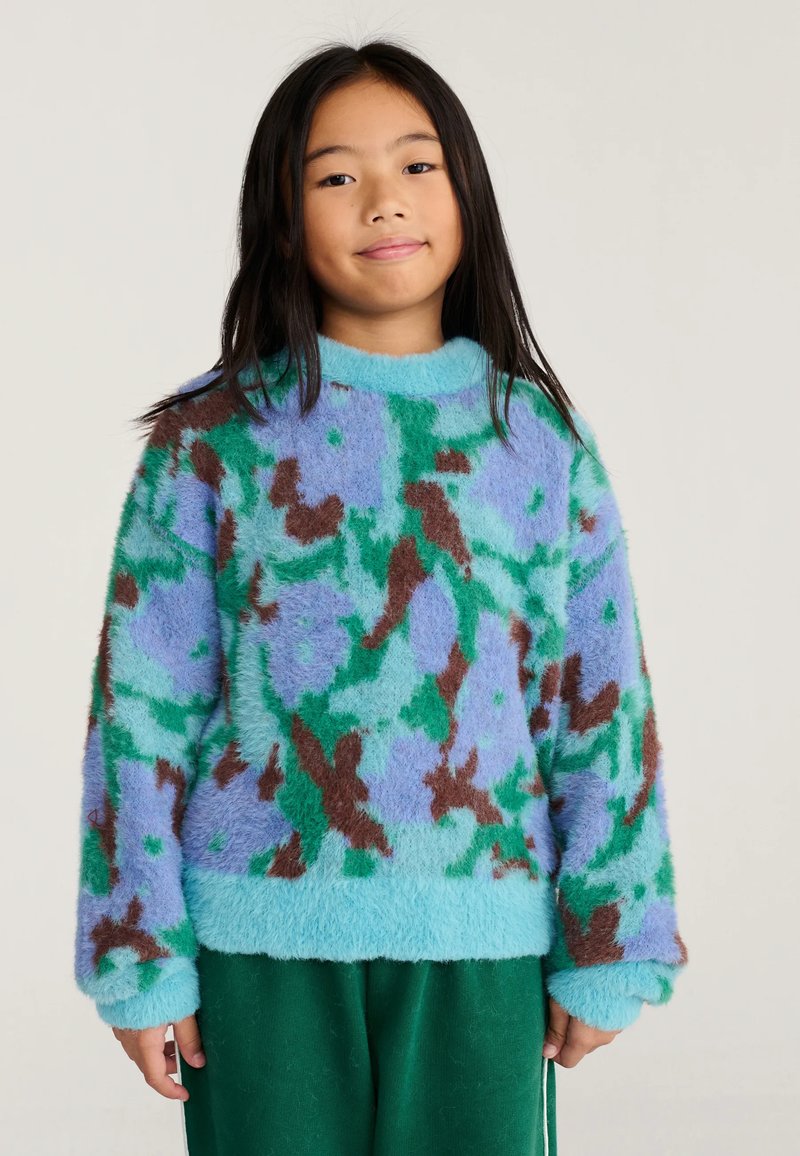 Jeune fille aux longs cheveux noirs portant un pull douillet avec un motif abstrait bleu, vert et marron et un pantalon vert foncé.