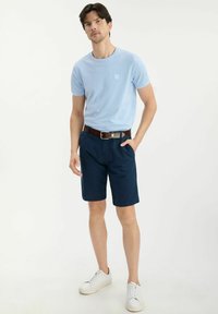 Jasnoniebieska koszulka z krótkim rękawem z małym logo, w zestawie z ciemnoniebieskimi szortami i białymi sneakersami, z brązowym paskiem.