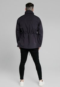 Mørk lilla jakke med snoreliv, lange ærmer og høj krave. Båret over sorte bukser og hvide sneakers. Minimalistisk design.