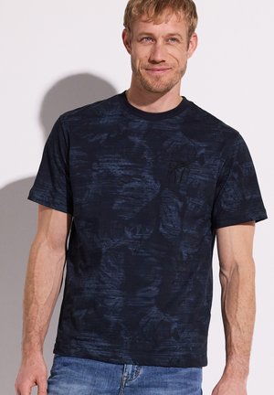 Homme souriant portant un t-shirt à manches courtes texturé bleu foncé et un jean bleu clair sur un fond clair uni.