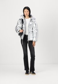 Chaqueta acolchada plateada con un acabado brillante, con capucha, cremallera frontal y puños elásticos, combinada con jeans negros de pierna acampanada y zapatillas negras.