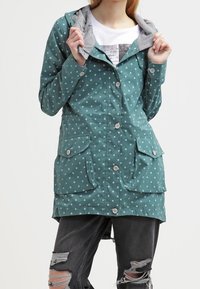 Morski płaszcz przeciwdeszczowy w białe kropki, z szarym kapturem, dwoma przednimi kieszeniami i zapięciem na guziki. Materiał wydaje się mieć fakturę i być wodoodporny.