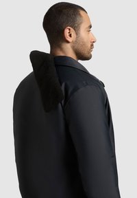 Mies, joka katsoo oikealle, pukeutuneena mustaan takkiin, jossa on pystykaulus, joka on vuorattu paksulla, tekstuurilla varustetulla mustalla kankaalla vaaleanharmaata taustaa vasten.