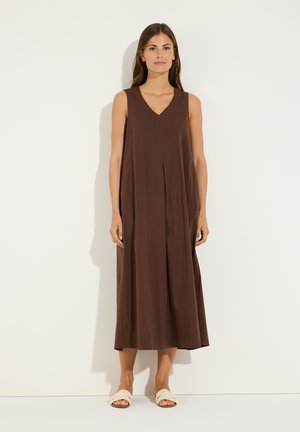 Frau, die ein ärmelloses braunes Midi-Kleid mit V-Ausschnitt und weiße flache Sandalen trägt, vor einem schlichten weißen Hintergrund.