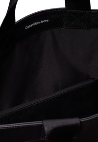 Musta olkalaukku, jossa on sileä sisäosa ja tekstuuri pohjaosa. Calvin Klein Jeans -brändäys valkoisena. Suuri ja tilava muotoilu.