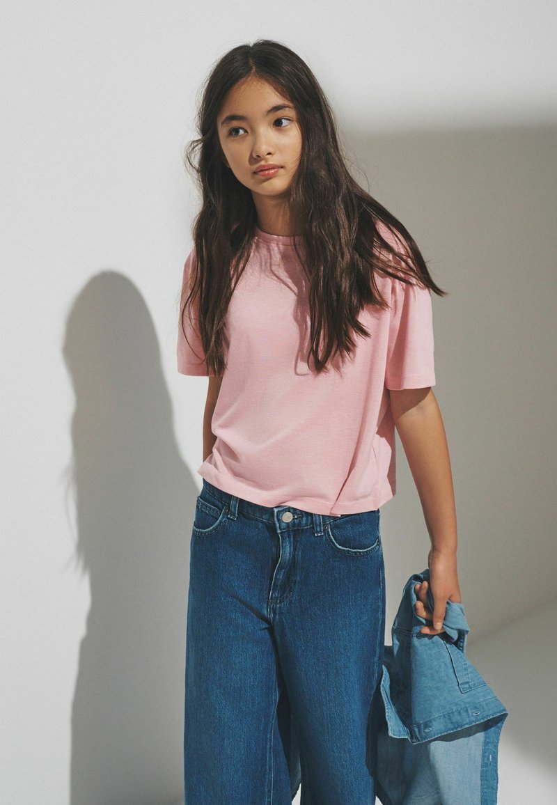Adolescente avec de longs cheveux foncés portant un t-shirt rose pâle et un jean bleu à jambes larges, tenant une veste en denim pliée dans sa main droite.