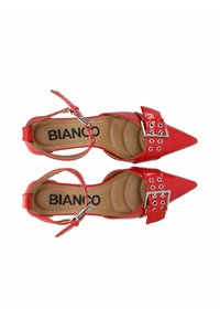 Tacones puntiagudos de charol rojo con tira al tobillo y detalle decorativo de hebilla. El interior cuenta con un suave acolchado y etiquetado de la marca.