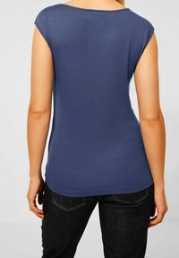 Tmavě modrý tričko s krátkým rukávem a kulatým výstřihem, hladká textura a přiléhavý střih. Nosené s tmavými džíny, ukazuje jednoduchý, ležérní design.