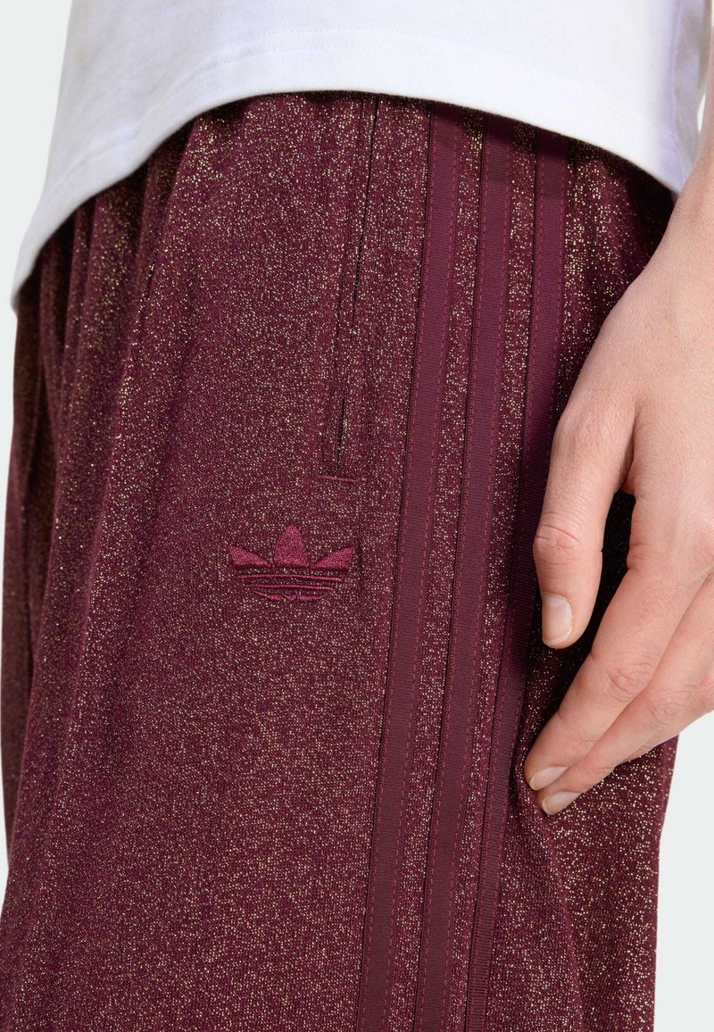 Nært billede af en hånd, der hviler på maroon Adidas træningsbukser med glitrende tekstur og det karakteristiske tre striber.