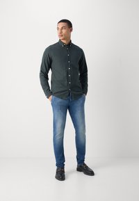 Dark green button-up shirt with a slim fit, paired with light blue jeans. Black shoes with gold accents complete the look. Simple background.