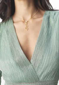 Femme portant une robe plissée verte à col en V et un collier en or avec de petits pendentifs ressemblant à des diamants sur sa poitrine.
