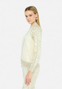 Giacca zip-up crema con maniche a motivo leopardo e dettagli dorati. Realizzata in materiale testurizzato, presenta tasche laterali e polsini a costine.