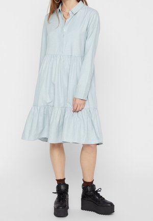 Kobieta ubrana w jasno-niebieską, długorękawową sukienkę z falbanami i kołnierzykiem, zestawioną z czarnymi botkami na platformie z sznurowadłami i ciemnymi skarpetkami do kostek.