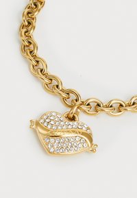 Kuldne ahelkäsir, millel on südame kujuline ripats, kaunistatud rhinestone'idega. Ripatsil on sileda pinna peal graveeritud "Guess".
