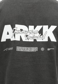 Temno siva majica s kapuco, ki vsebuje belo besedilo z blagovno znamko "ARKK" in frazo "Inovacija vključena," ter manjše tiskane podrobnosti in logotipe.