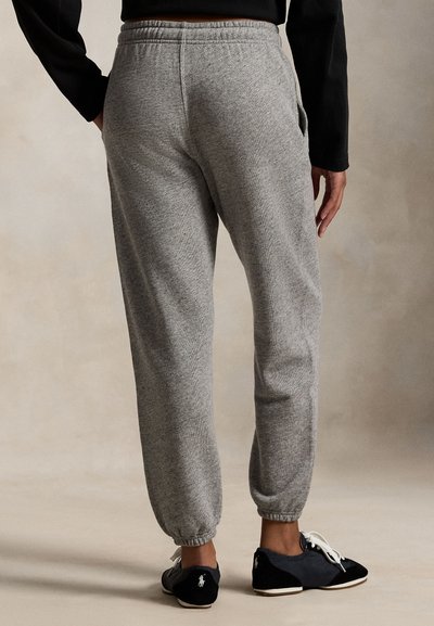 Pelēkas sporta bikses ar elastīgām aprocēm un jostas daļu, ar mīkstu tekstūru un sānu kabatām, kopā ar melniem un zilajiem apaviem.