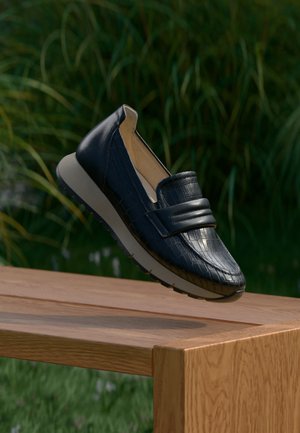 Schwarzer Slip-On-Loafer mit strukturiertem Leder und gepolstertem Riemen, der über einer Holzbank im Freien mit grünem Gras im Hintergrund schwebt.